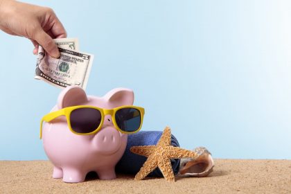 Saving for beach vacation or retirement, with pink piggy bank and sunglasses.  Studio shot with plain blue background.  Sharp focus on the five dollar bill.  Space for copy.  Warm color and directional lighting are intentional.